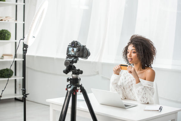 selectieve focus van aantrekkelijke Afrikaanse Amerikaanse influencer houden credit card in de buurt van laptop en kijken naar digitale camera  - Foto, afbeelding