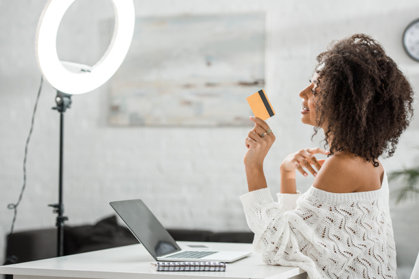 visão lateral de influenciador americano africano feliz em aparelho segurando cartão de crédito perto de laptop
  - Foto, Imagem