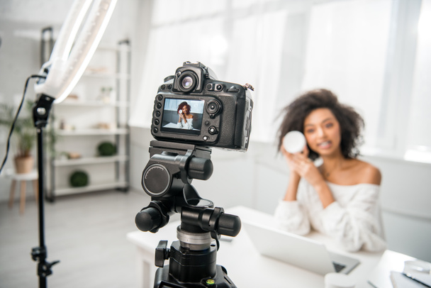selective focus of digital camera with attractive african american influencer in braces holding container with cosmetic cream on display  - 写真・画像