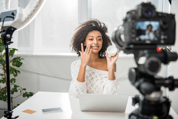 messa a fuoco selettiva di felice africano americano video blogger in bretelle applicando crema cosmetica vicino fotocamera digitale
  - Foto, immagini