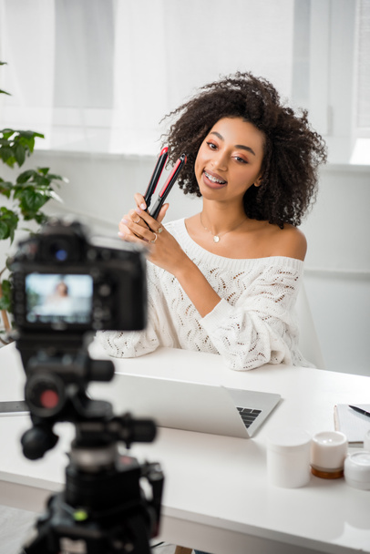 selective focus of happy african american influencer in braces holding hair straightener near laptop and digital camera  - Photo, Image
