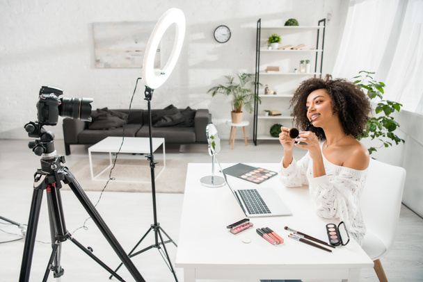 heureux blogueur vidéo afro-américain dans bretelles tenant pinceau cosmétique près de l'appareil photo numérique
  - Photo, image