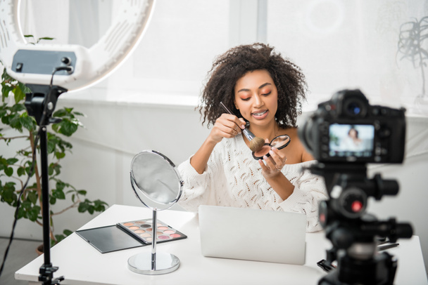 messa a fuoco selettiva di felice africano americano video blogger in bretelle che tengono pennello cosmetico vicino alla fotocamera digitale
  - Foto, immagini