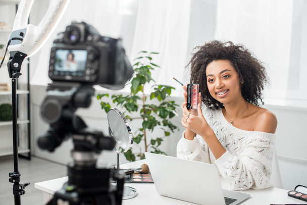 selective focus of happy african american video blogger in braces holding palette with lip gloss near digital camera  - Zdjęcie, obraz