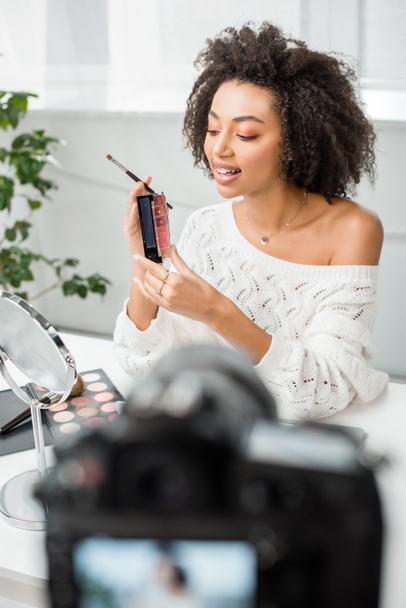 selectieve focus van gelukkig Afrikaans Amerikaans video blogger in beugels met palet met lip gloss in de buurt van digitale camera  - Foto, afbeelding