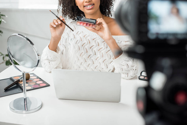 vue recadrée du blogueur vidéo afro-américain heureux dans des bretelles tenant la palette avec du brillant à lèvres près de l'appareil photo numérique
  - Photo, image