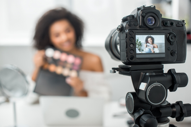 mise au point sélective de l'appareil photo numérique avec heureuse fille afro-américaine dans des bretelles tenant palette avec ombre à paupières et pinceau cosmétique sur l'affichage
  - Photo, image