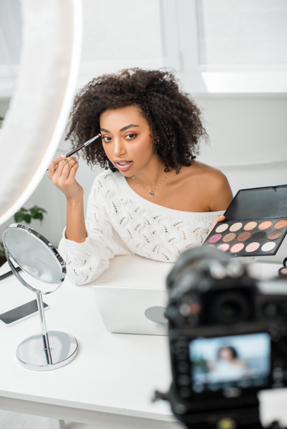 selective focus of beautiful african american video blogger applying eye shadow and looking at digital camera  - Photo, Image