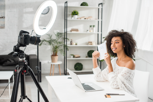 happy african american video blogger in braces holding notebook with influencer marketing lettering near digital camera  - Fotó, kép