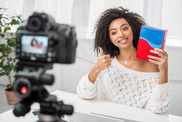 foyer sélectif de l'influenceur afro-américain heureux dans les bretelles tenant boîte colorée et montrant pouce près de l'appareil photo numérique
  - Photo, image