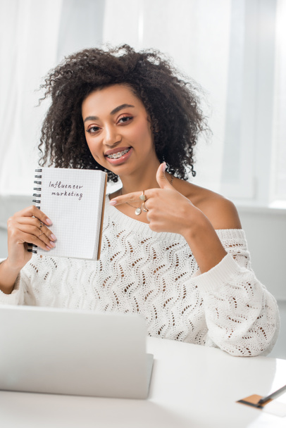 alegre afroamericana chica en frenos apuntando con el dedo en el cuaderno con influencer marketing letras en casa
 - Foto, imagen