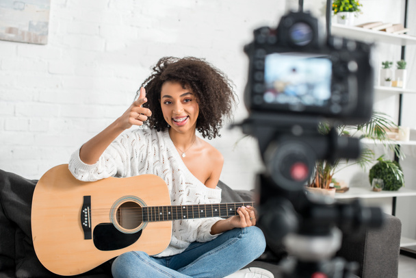 selektiver Fokus eines fröhlichen afrikanisch-amerikanischen Mädchens in Hosenträgern, das eine akustische Gitarre hält und mit dem Finger auf die Digitalkamera zeigt  - Foto, Bild