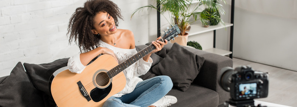 panoramic shot of cheerful african american girl in braces holding acoustic guitar and looking at digital camera  - Fotó, kép