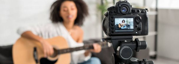 panoramic shot of digital camera with young african american girl in braces playing acoustic guitar and singing on display  - Photo, Image