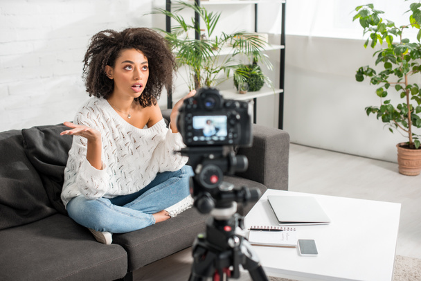 selective focus of curly african american influencer in braces gesturing and talking near digital camera in living room  - Fotografie, Obrázek