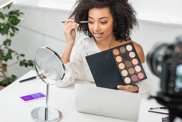 selective focus of cheerful african american video blogger in braces applying eye shadow near smartphone with shopping app and digital camera  - Photo, Image