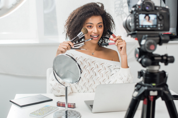 szelektív fókusz boldog afro-amerikai befolyásoló fogszabályzó tartja ajakfények és kozmetikai kefék közelében okostelefon online térkép a képernyőn és a digitális fényképezőgép - Fotó, kép