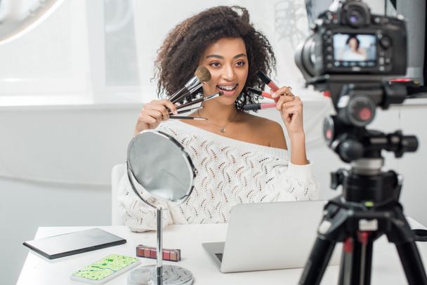 selective focus of happy african american influencer in braces holding lip glosses and cosmetic brushes near smartphone with map on screen and digital camera - Photo, Image