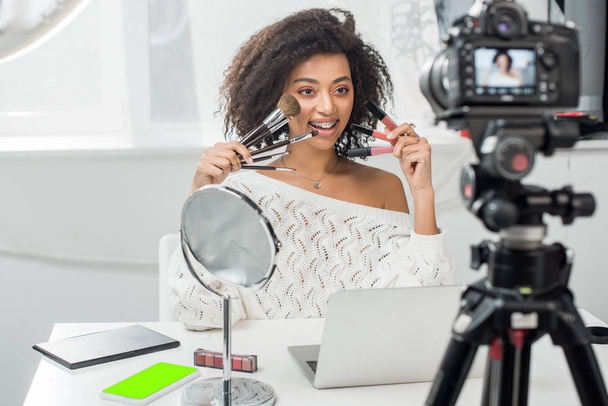 επιλεκτική εστίαση της χαρούμενης αφρικανικής αμερικανική επιρροή σε τιράντες κρατώντας lip gloss και καλλυντικά πινέλα κοντά smartphone με πράσινη οθόνη και ψηφιακή φωτογραφική μηχανή - Φωτογραφία, εικόνα