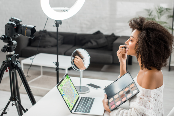 vue latérale d'un heureux influenceur afro-américain tenant une palette d'ombres à paupières près d'un ordinateur portable avec carte en ligne à l'écran et appareil photo numérique
  - Photo, image