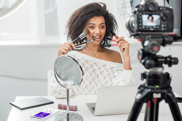 KYIV, UKRAINE - DECEMBER 10, 2019: selective focus of african american girl in braces holding lip glosses and cosmetic brushes near smartphone with music app  - 写真・画像