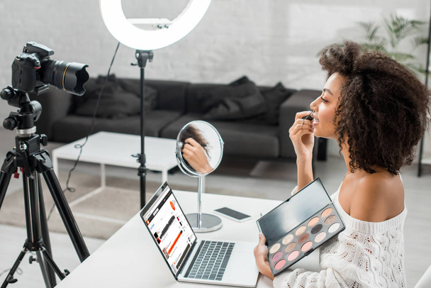 KYIV, UKRAINE - DECEMBER 10, 2019: side view of african american influencer holding eye shadow palette near laptop with soundcloud website and digital camera  - Фото, зображення