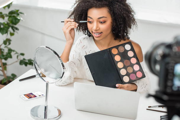 KYIV, UKRAINE - DECEMBER 10, 2019: selective focus of african american influencer in braces applying eye shadow near smartphone with soundcloud app on screen  - Photo, image