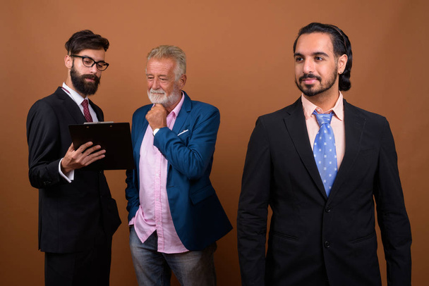 Kolme monietnistä parrakasta liikemiestä yhdessä ruskeaa taustaa vasten
 - Valokuva, kuva