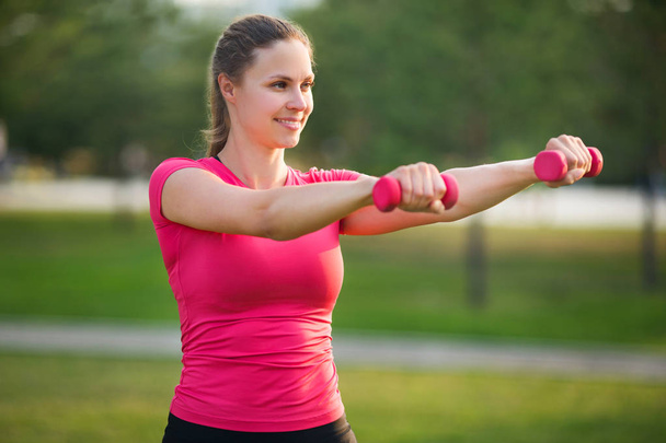 beautiful young woman in sportswear is engaged in training with dumbbells in the summer in the park - Foto, imagen