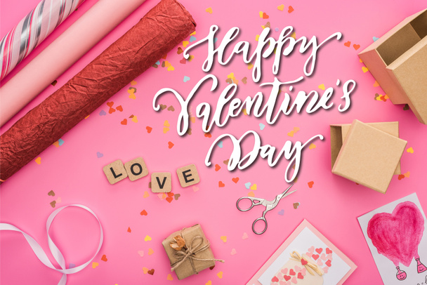 top view of valentines confetti, scissors, wrapping paper, gift boxes, greeting cards and love lettering on wooden cubes on pink background with happy valentines day illustration - Fotó, kép