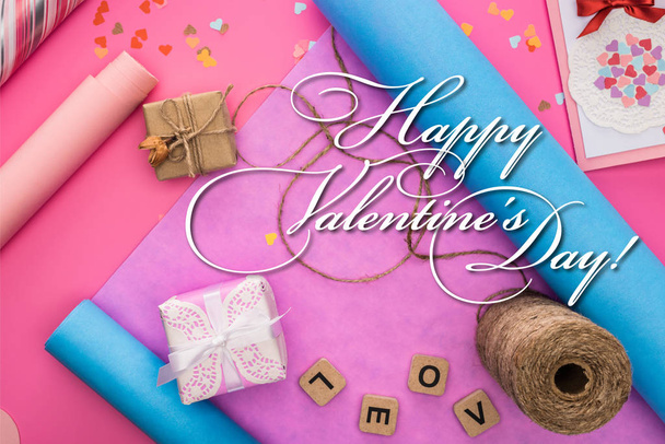 vrchní pohled na valentinky dekorace, balicí papír, motouzy, dárkové krabice, přání a láska nápisy na dřevěné kostky na růžovém pozadí s šťastným Valentýna ilustrace - Fotografie, Obrázek