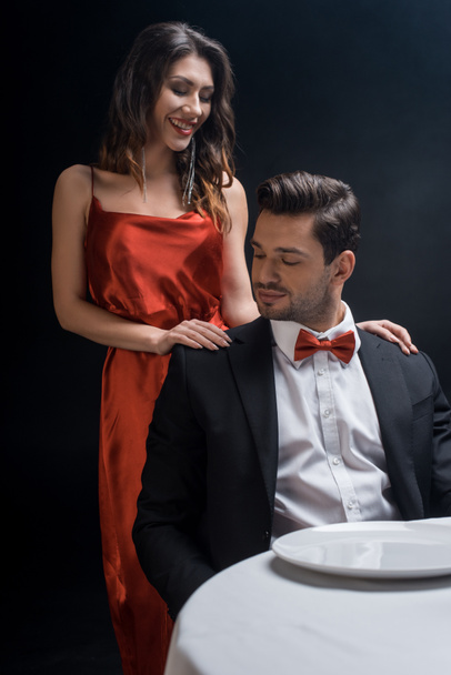 Smiling woman in elegant dress standing by handsome man at served table isolated on black - Foto, Bild