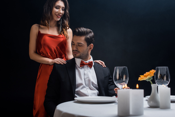 Mulher sorridente bonita de pé ao lado de homem bonito em terno na mesa servida isolado em preto
 - Foto, Imagem