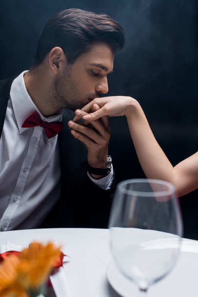 Side view of man in suit kissing hand of woman at served table on black background with smoke - Fotoğraf, Görsel