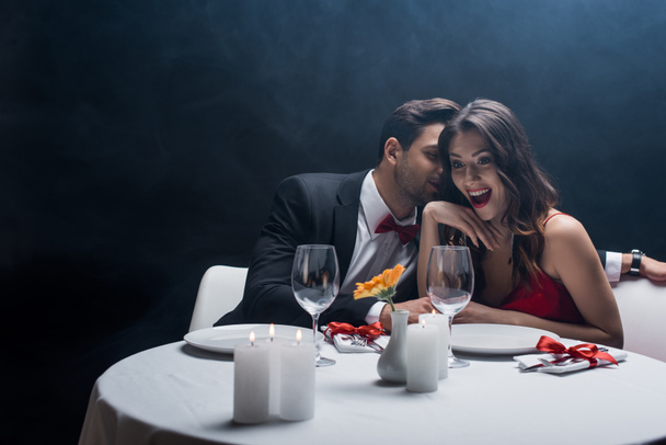 Handsome man in suit whispering to shocked woman at served table on black background with smoke - Photo, Image