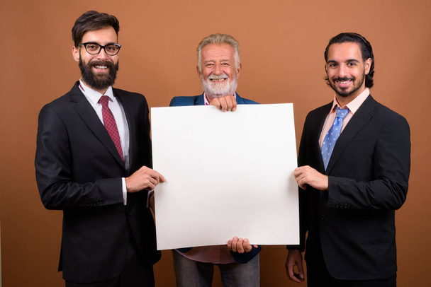 Kolme monietnistä parrakasta liikemiestä yhdessä ruskeaa taustaa vasten
 - Valokuva, kuva