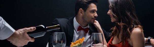 Focus selettivo di coppia sorridente a tavola accanto al cameriere versando vino in bicchieri isolati su nero
 - Foto, immagini