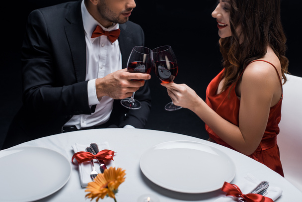 Boční pohled na elegance pár cinkání s červeným vínem u servírovaného stolu izolované na černé - Fotografie, Obrázek