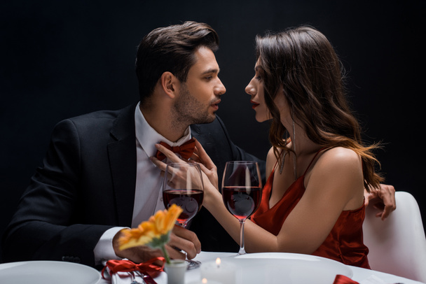 Side view of beautiful woman adjusting bow tie of handsome man during romantic dinner isolated on black - Fotó, kép