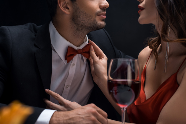 Cropped view of elegant woman adjusting bow tie of man in suit by wine glass isolated on black - Photo, Image