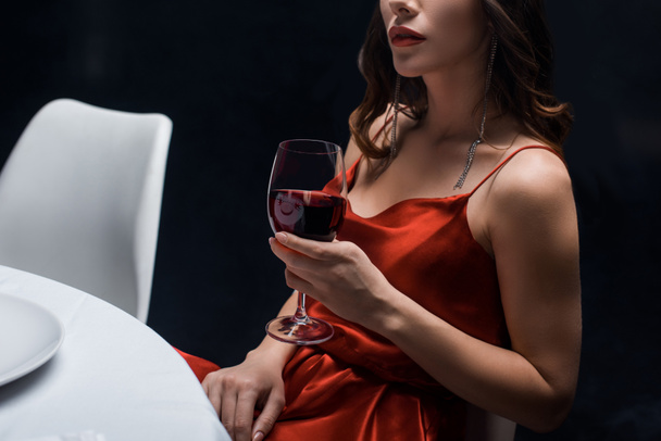 Vista recortada de mujer elegante en vestido rojo sosteniendo copa de vino en la mesa aislada en negro
 - Foto, Imagen