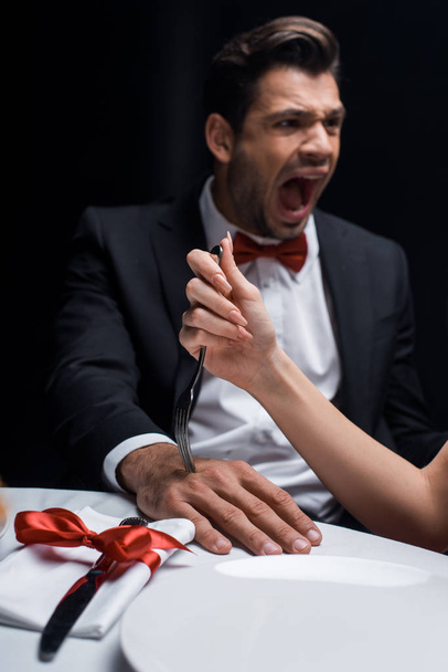 Selective focus of woman sticking fork in hand of screaming boyfriend during romantic dinner isolated on black - Photo, Image