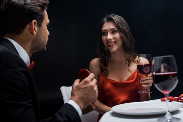 Handsome man presenting jewelry box at smiling girlfriend during romantic dinner isolated on black - Photo, Image