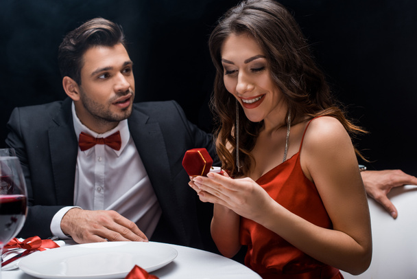 Όμορφος άντρας κοιτάζει χαμογελαστή φίλη με δαχτυλίδι κοσμημάτων κατά τη διάρκεια ρομαντικού δείπνου απομονωμένο σε μαύρο - Φωτογραφία, εικόνα
