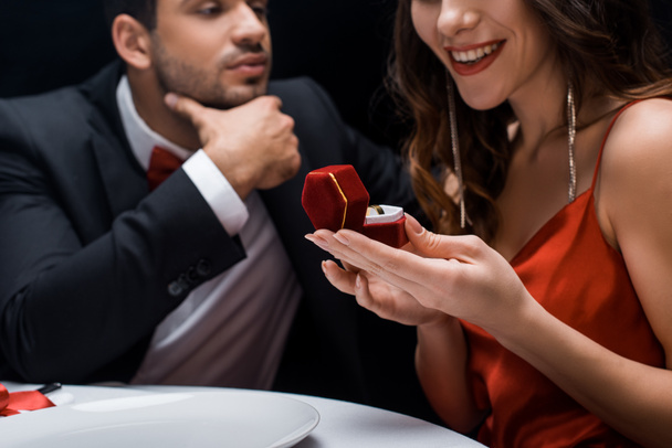 Selective focus of smiling woman holding jewelry ring in box by handsome boyfriend at served table isolated on black - Fotoğraf, Görsel