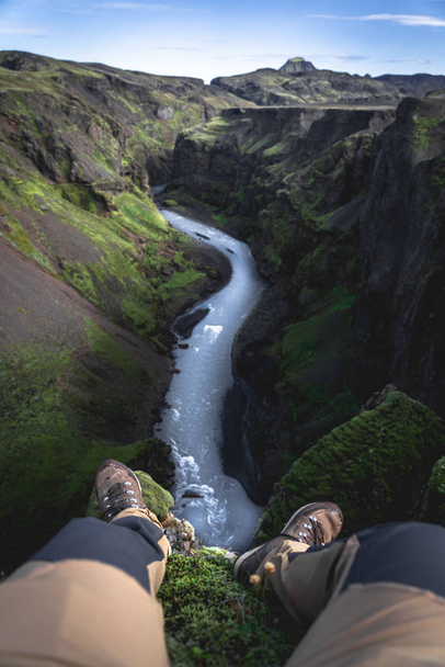 Pernas penduradas no penhasco sobre o rio em um desfiladeiro durante a aventura nas terras altas da Islândia
 - Foto, Imagem