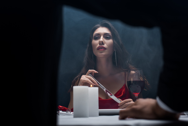 Selective focus of beautiful girl holding knife and looking at boyfriend during romantic dinner on black background with smoke - Fotografie, Obrázek