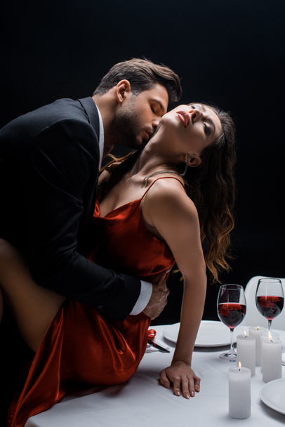 Side view of handsome man kissing beautiful woman in neck on served table isolated on black - Photo, Image