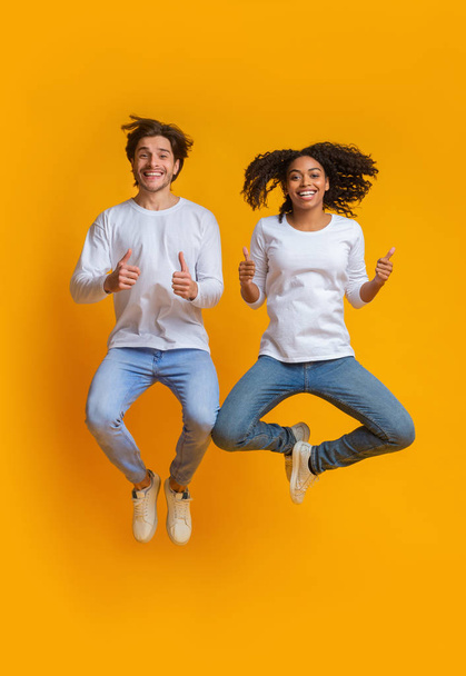 Joyful interracial couple jumping in the air and gesturing thumbs up - Foto, Bild