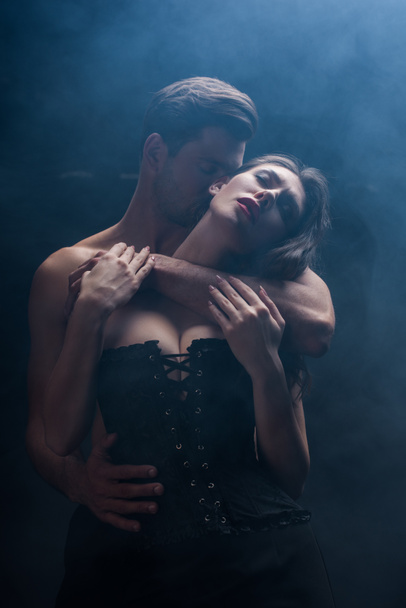 Handsome man kissing and hugging seductive woman in corset on black background with smoke - Photo, Image